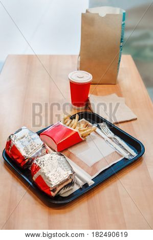 Closeup shot image of two burgers wrapped in tinfoil french fries potato chips red cup of coffee or tea on tray on wooden table in fast food restaurant