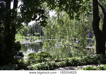 Laval, Quebec - June 14, 2015 - Landscape of people walking on paths alongside the small lake in the Nature Park, Laval, Quebec framed by foliage in the foreground on a sunny day in June.