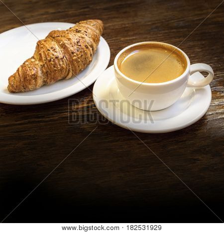 A square photo of cup of dark coffee with cereal croissant