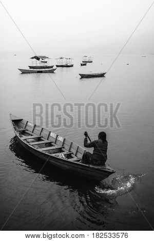 A woman alone in a canoe boat paddling on Fewa Lake Pokhara on a misty morning in black and white. Pokhara Nepal.