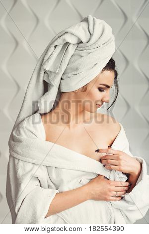 sexy young girl with dark hair, big eyes and dark eyebrows wearing white bath robe whith towel on her head. Model with light nude make-up, white studio background, beauty photo