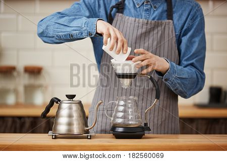 Closeup of barista in uniform pouring ground coffee from white paper cup making pourover coffee.