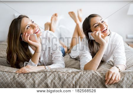 Two beautiful girls talking on the phone and smiling while lying on a luxorious bed