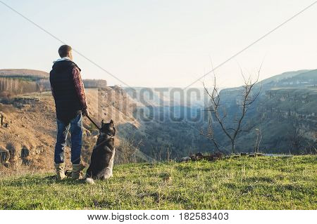 A man with a beard walking his dog in the nature, standing with a backlight at the rising sun, casting a warm glow and long shadows against the background of the gorge and trees on caucazus