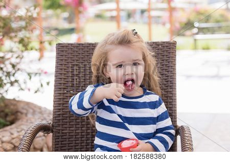 food and restaurant. cute baby boy small little child with long blond hair in blue striped clothes eating tasty ice cream frozen pink dessert in cup on chair in outdoor cafe on sunny day