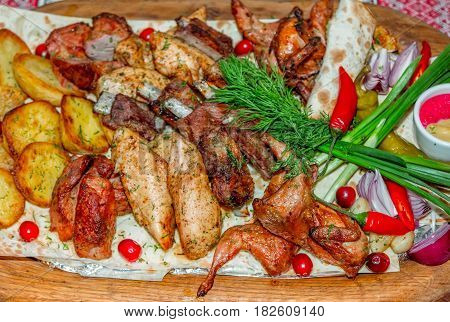 Assorted barbeсue plate. Grilled potato and meat platter- beef, pork ribs, quails, chicken fillet served with flatbread, chili pepper, red onion, dill, horseradish and mustard on a large wooden tray.