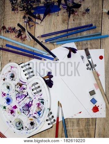 A blank sheet of paper for text surrounded by dried flower petals on a wooden background. Illustration of a hobby, drawing and inspiration. Top view.Easel, brushes, watercolor paint