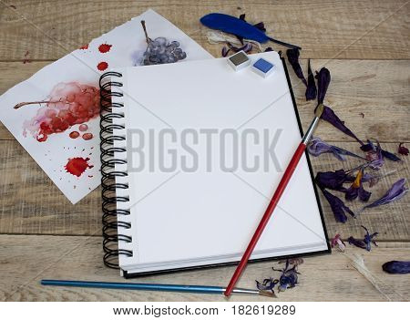 A blank sheet of paper for text surrounded by dried flower petals on a wooden background. Illustration of a hobby drawing and inspiration. Top view.Easel brushes watercolor paint