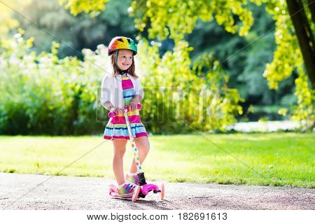 Little child learning to ride a scooter in a city park on sunny summer day. Cute preschooler girl in safety helmet riding a roller. Kids play outdoors. Active leisure and outdoor sport for children.