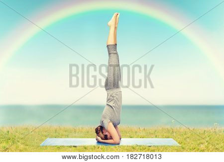 fitness, sport, people and healthy lifestyle concept - woman making yoga in headstand pose on mat over blue sky, rainbow and sea background