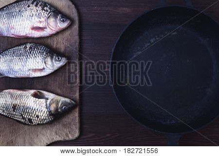 Three live carp fish in a scales on a kitchen board next to a black cast-iron frying pan