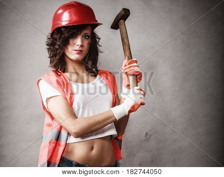 Sex equality and feminism. Sexy girl in workwear safety helmet and orange vest holding hammer tool on gray. Young attractive woman working as construction worker. Studio shot.
