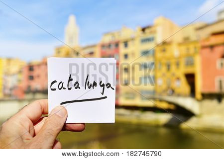 closeup of the hand of a young man holding a paper note with the text Catalunya written in it in Girona, Spain, with the Onyar River and some of the characteristics colorful houses of its Old Town