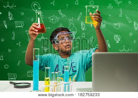 Kids and Science concept - Cute Indian little boy busy doing  science or chemistry experiment with test tube and flask with safety eye glass over green chalkboard background with science doodles drawn