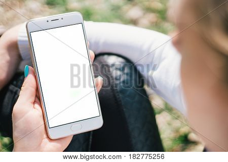 Girl with smartphone in hands with mock up green screen of blank screen sits in park on open space, screen for content integration. Hands holding gadget on blurred backdrop, front