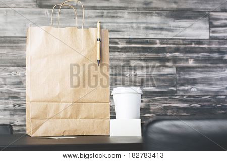 Front view of brown shopping bag with pen and coffee cup with empty business card on wooden background. Mock up