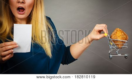 Shocked woman holding shopping basket with bread looking at bill receipt being scared of huge prices