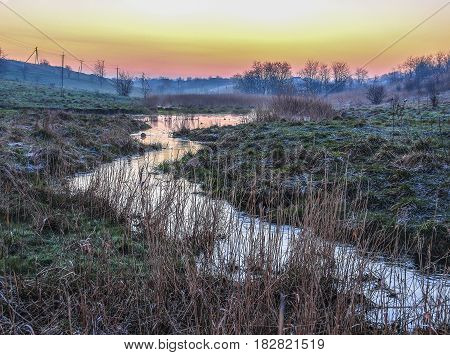 The stream in the hollow in March in the Krasnodar region. Orange sunrise in the sky, and the grass is covered with white bloom from the morning frost.