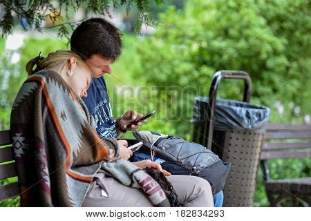 RIGA LATVIA - JULY 27 2016: Unknown young couple sitting on a park bench and in smartphone play Pokemon Go.