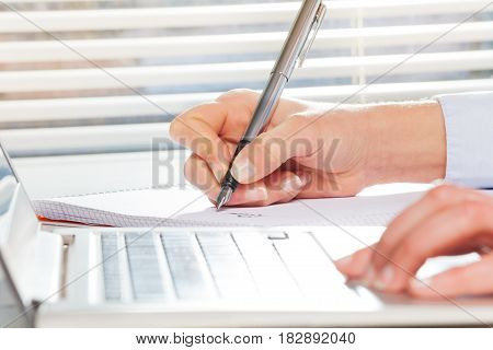Woman's hands writing on notebook while working on the computer next to the window