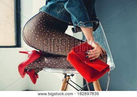 gorgeous girl dressed in a blue jeance jacket, red high heel shoes with red small bag and transparent tights without panties. posing sitting on a stylish armchair. bare shoulders, view from behind