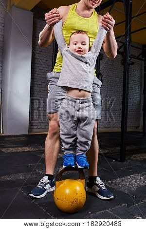 Happy father holds the hands of his son who stands on the kettlebells against brick wall in the cross fit gym. Sporty father and lettle son in the sportwear and sportive shoes. Rearing child of healthy lifestyle. Cross fit concept.