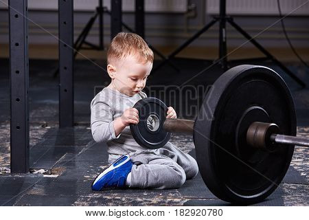 Little cute child in sportwear in the cross fit gym with a barbell. Little boy in the blue sporty shoes. Portrait of sporty child. Rearing child in the healthy lifestyle. Concept of active life for children.