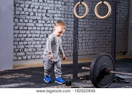 Little cute boy in the grey sportwera standing near barbell against brick wall in the cross fit gym. Sporty child in the blue sportive shoes. Rearing in the healthy lifestyle. Concept of the active child's life.