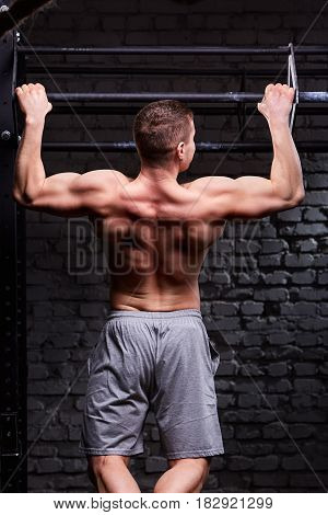 Rear view photo of muscular male doing exercises on horizontal bar against brick wall at the cross fit gym. Sportsman in the grey sportive shorts. Body and muscle. Power and energy. Healthy lifestyle.