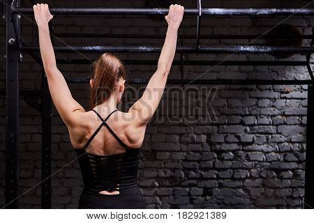 Rear view photo of young muscular woman doing exercises on horizontal bar against brick wall at the cross fit gym. Body and muscle. Power and energy. Healthy lifestyle.
