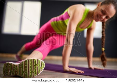Beautiful girl doing yoga exercises on the violet yoga mat at the gym against blue background. Sporty woman in the sportwear, green t-shirt, pink leggings and sportive shoes. Healthy lifestyle.