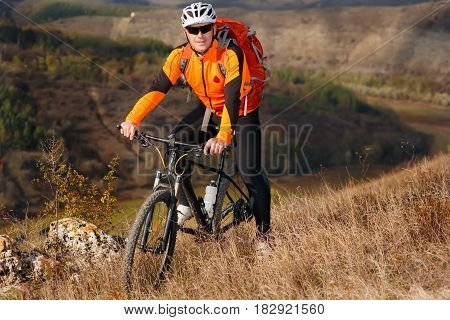 Cyclist in the orange jacket standing with his bike on the top of a hill against beautiful landscape. Hill and rock in the countryside. Sportsman in the helemet, sunglasses and with red backpack. Extreme travel in the summer season.