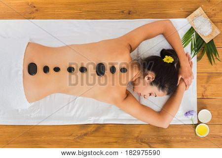 Black marble stone massage in spa. Top view of beautiful girl with stones on back and rose petals in wellness center. Pretty woman relaxation procedure, attractive young indian girl in beauty parlor.