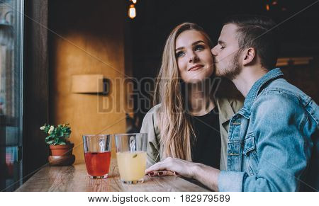 Portrait of beautiful young couple in love having great time at nice coffee shop. Attractive guy with a beard kissing his gorgeous girlfriend in a cheek. Close up