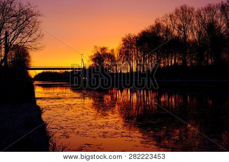 Dramatic And Colorful Sunrise Over A Beautiful Early Winter Landscape With A Frozen River Or Canal, 