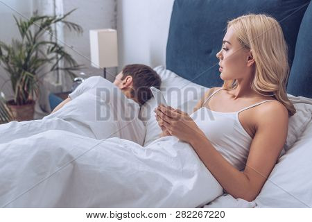 Jealous Young Woman Using Smartphone And Looking At Boyfriend Sleeping In Bed, Distrust Concept