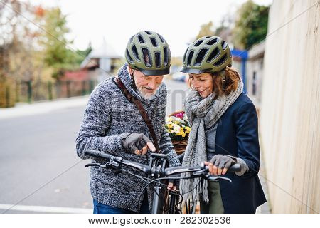 Active Senior Couple With Electrobikes Standing Outdoors On Pathway In Town.