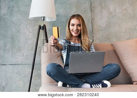 Beautiful Woman Showing Credit Card And Sitting On Sofa With Laptop