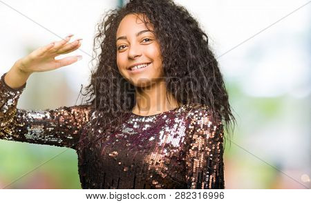 Young beautiful girl with curly hair wearing night party dress gesturing with hands showing big and large size sign, measure symbol. Smiling looking at the camera. Measuring concept.