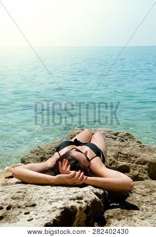 Back View Young Woman In Black Swimsuit And Straw Hat On Wild Snone Beach With Clean Sea.