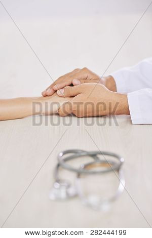 Hands Of Young Doctor In Whitecoat And Those Of His Patient On Desk With Stethoscope Near By