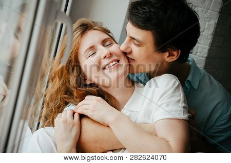 Happy Girl And Man Kissing Near Window In Home. White And Blue Clothes. Valentine Day.
