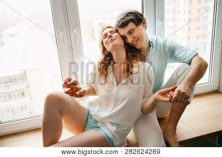Happy Girl And Man Hugging Near Window In Home. White And Blue Clothes. Valentine Day.