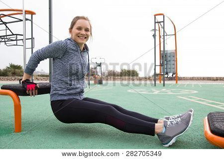 Joyful Sporty Girl Working On Arm Muscles. Young Woman In Hoody And Tights Doing Triceps Bench Dips 