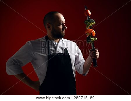Bearded Professional Chef In Uniform Going To Fight With Skewer With Vegetables On It. Stands Like A