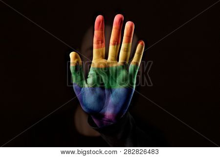 closeup of a young caucasian person with the palm of his or her hand, patterned with a rainbow flag, in front of his or her face, against a black background