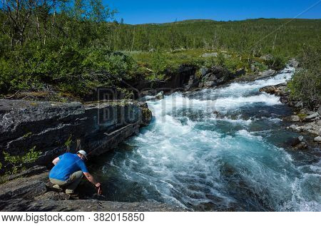 Summer trekking in Sweden. Man fills bottle with clear drinking water from river Abiskojokk in Abisko National Park in northern Sweden, Scandinavian nature. Sunny day