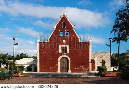 Front Of The Red Colonial Church Itzimna In A Sunny Park With Trees, Merida, Yucatan, Mexico