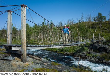 Hiking in Sweden in summer. Man crossing river on wooden suspension bridge, Trekking in Abisko National Park in Lapland. Arctic nature of Scandinavia in warm sunny day