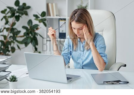 Young tired businesswoman with headache touching temple and bending slightly forwards over desk while trying to concentrate on work